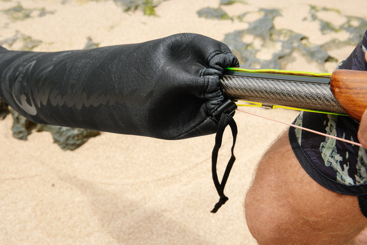 Close-up of a black Waihana-branded neoprene speargun sock being fitted over a carbon fiber speargun barrel with yellow bungee bands, with sandy beach and rocks in the background.