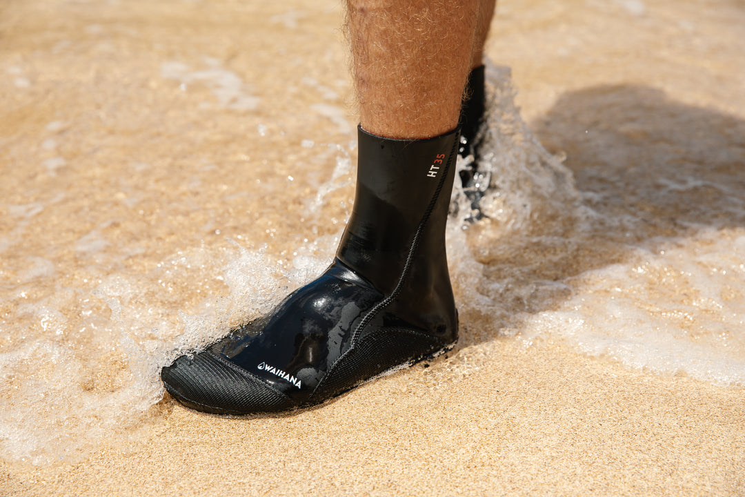 Close-up of a man wearing black Waihana HT35 neoprene dive socks standing in the shallow shore wash on a sandy beach.