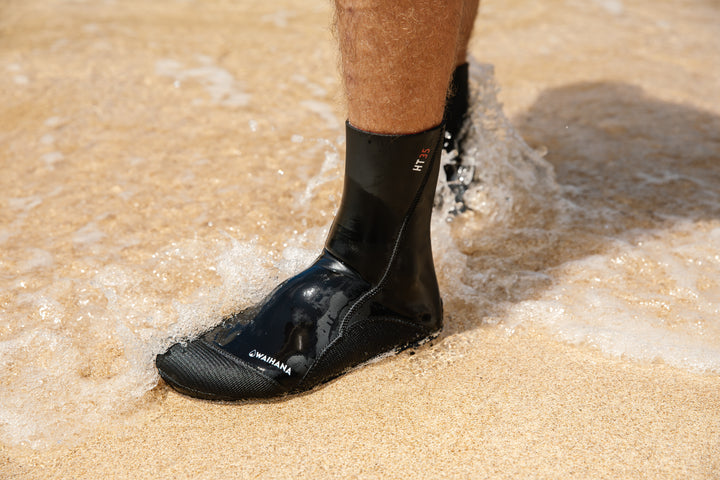 Close-up of a man wearing black Waihana HT35 neoprene dive socks standing in the shallow shore wash on a sandy beach.