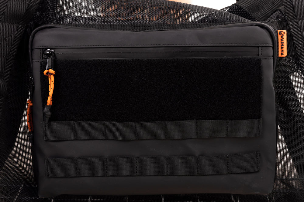 Close-up of a black Waihana gear bag front pocket with MOLLE webbing straps, velcro patch panel, zipper with orange paracord pull, and an orange Waihana logo tag in the corner.