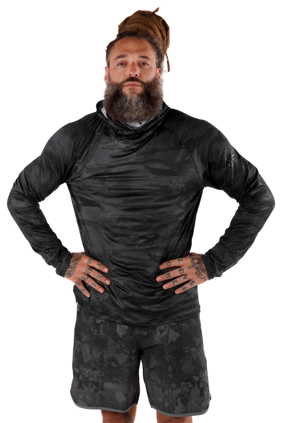 Male model wearing the Waihana Tropicam Black long-sleeve sun shirt with integrated neck gaiter in a dark camouflage pattern, paired with matching camo shorts, hands on hips against a white studio background.