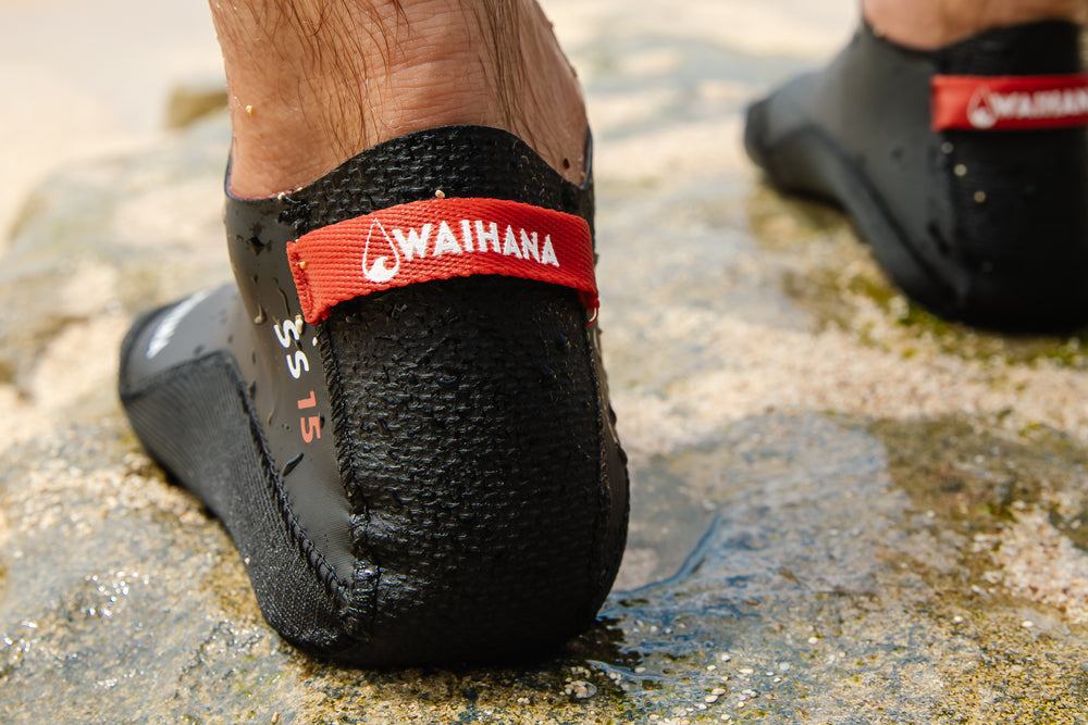 Close-up of a foot wearing a black Waihana neoprene dive bootie with a red branded strap, standing on a wet rocky surface near water.